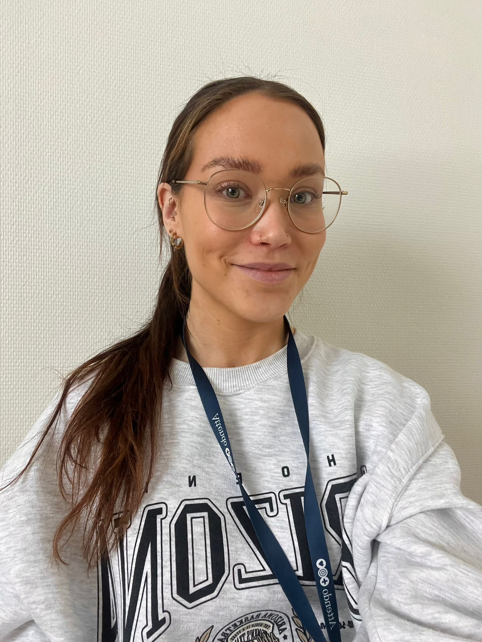 Nainen hymyilee kameraan pukeutuneena harmaaseen collegepaitaan ja silmälaseihin valkoista taustaa vasten