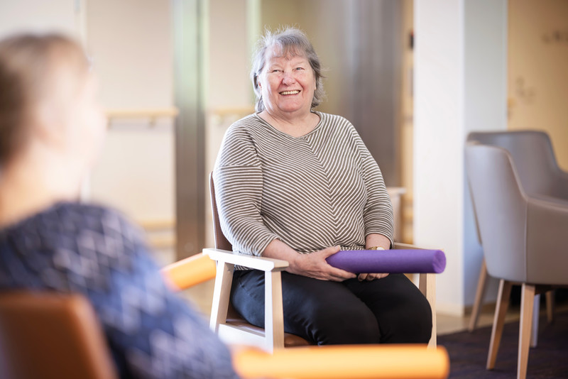 Iäkäs nainen osallistuu jumppatuokioon istuen tuolilla ja hymyillen. Hän pitelee violettia vaahtorullaa. 