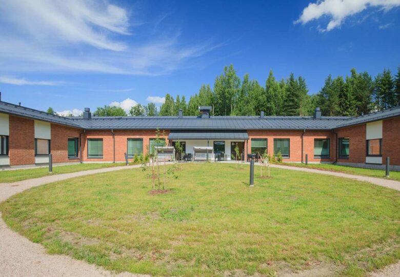 Yksikerroksinen Hiidenhovin punertava rakennus ulkoa, etualalla nurmikkoa