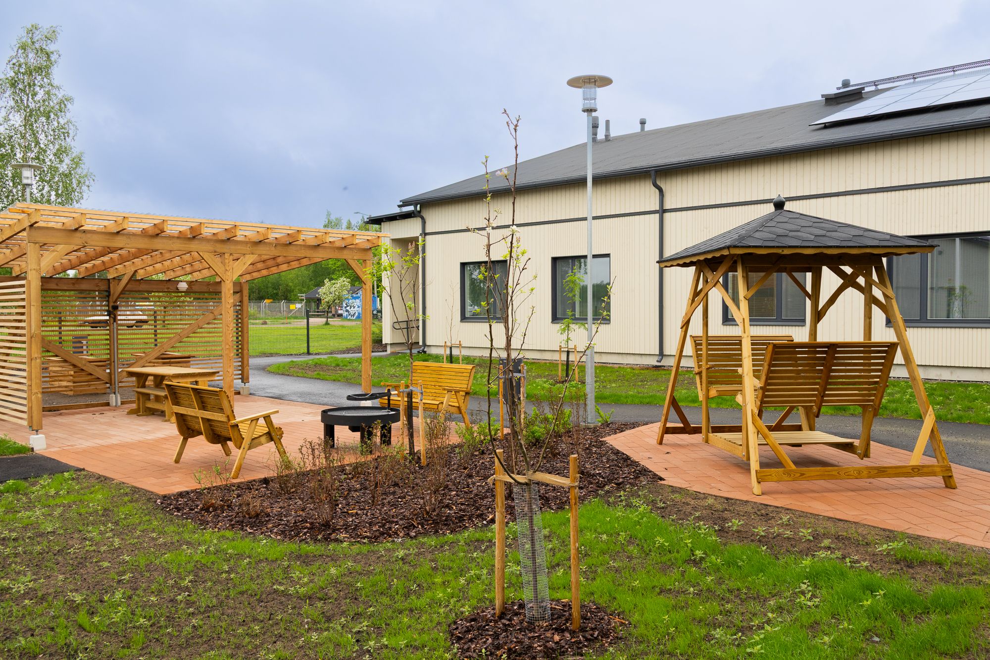 Pilotintuvan puutarhaa, pergola ja keinu, taustalla vaalea yksikerroksinen hoivakotirakennus