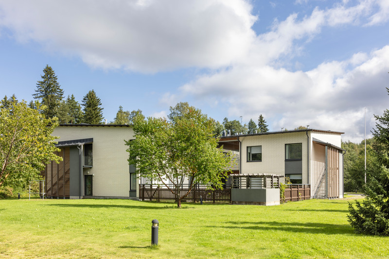 Attendo Villa Aallon hoivakoti kuvattuna pihan puolelta kesäisenä päivänä. Vaaleat rakennukset ja puinen terassi sijaitsevat vehreällä nurmialueella, ja taustalla näkyy korkeita puita sinistä taivasta vasten.