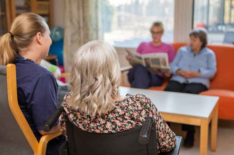 Neljä henkilöä hoivakodin olohuoneessa istumassa ja lukemassa sanomalehteä