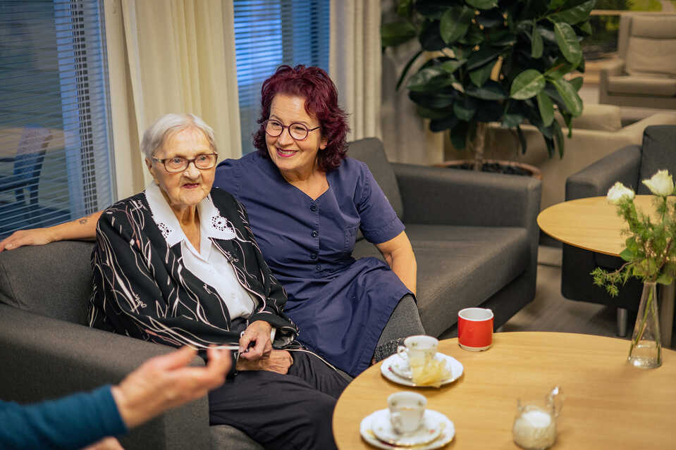 hoitaja ja hoivakodin asukas istuvat vierekkäin sohvalla, pöydällä kahvikupit