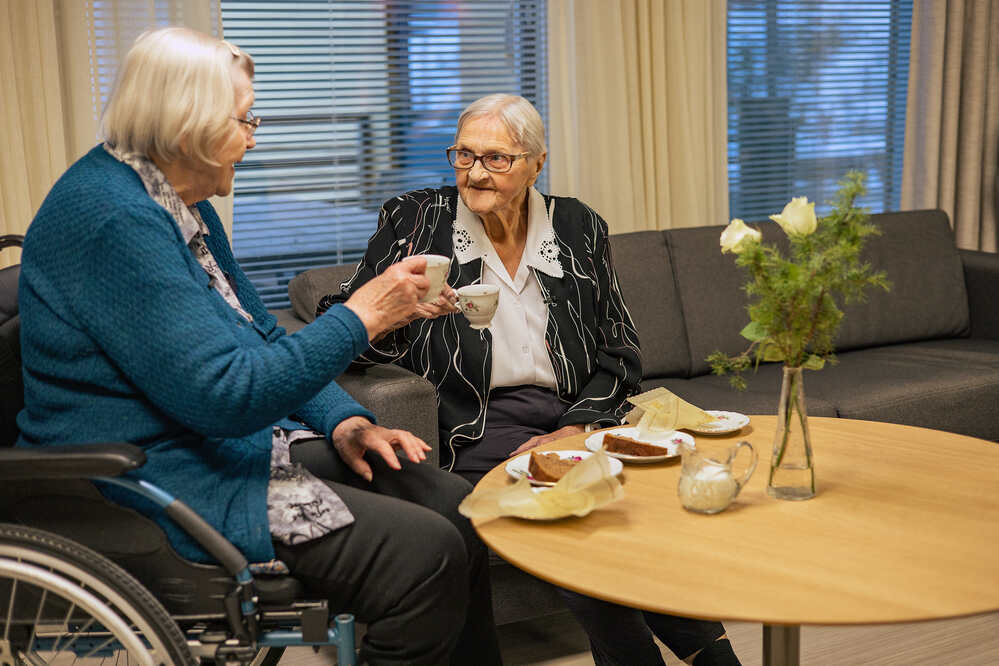 Kaksi vanhempaa naista nauttimassa kahvia ja kakkua olohuoneessa, kukkamaljakko pöydällä