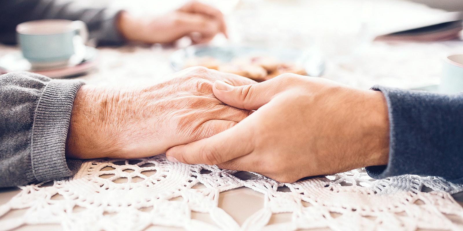 vanhemman ja nuoremman kädet käsi kädessä pöydällä, jossa on pitsiliina