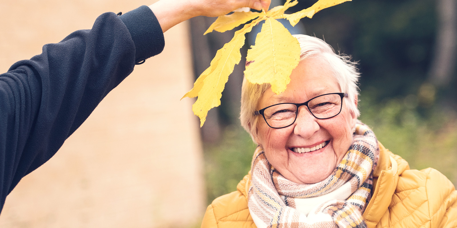 Iäkäs nainen hymyilee syystakkissa, vaahteranlehdet pään päällä, syksyinen tausta