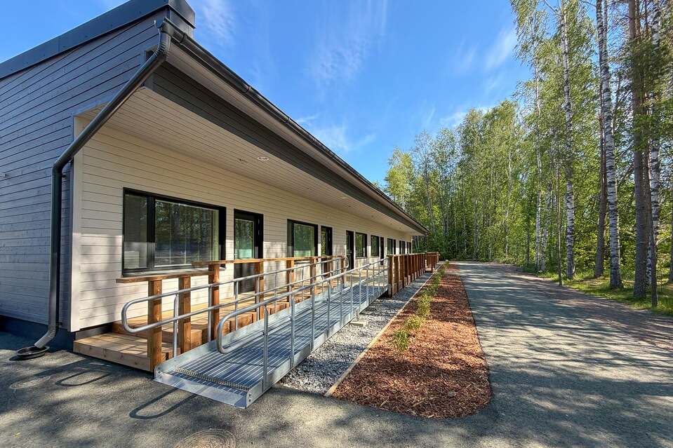 Kotojärven yksikerroksinen vaalea rakennus, etualalla pyörätuoliluiska