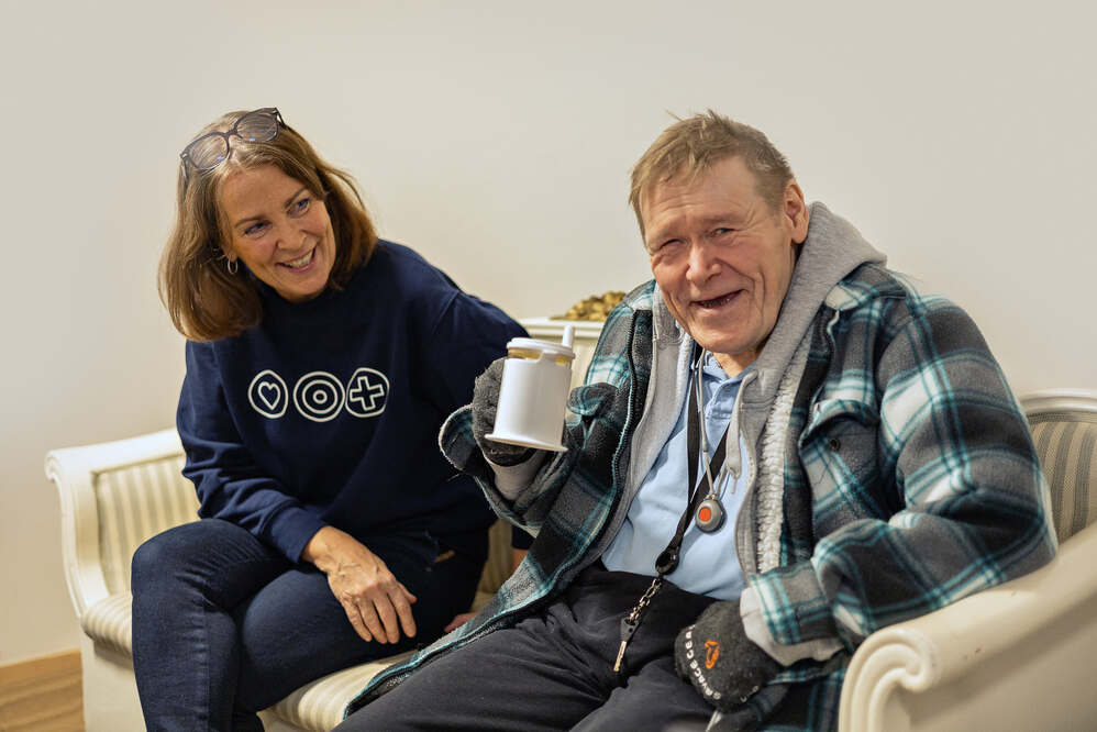 Nainen ja mies istuvat sohvalla, mies hymyilee ja pitää mukia kädessään.