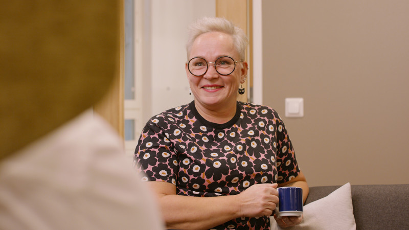 vaaleatukkainen nainen istuu sohvalla kahvikuppi kädessä ja hymyilee