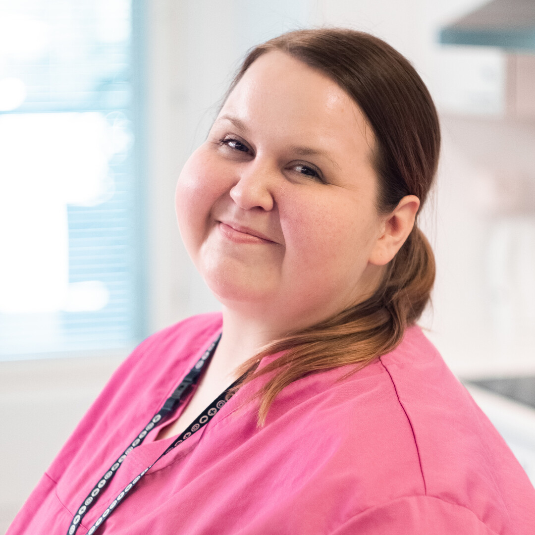 Lähihoitaja hymyilee kameralle, vaaleanpunainen työvaate päällään