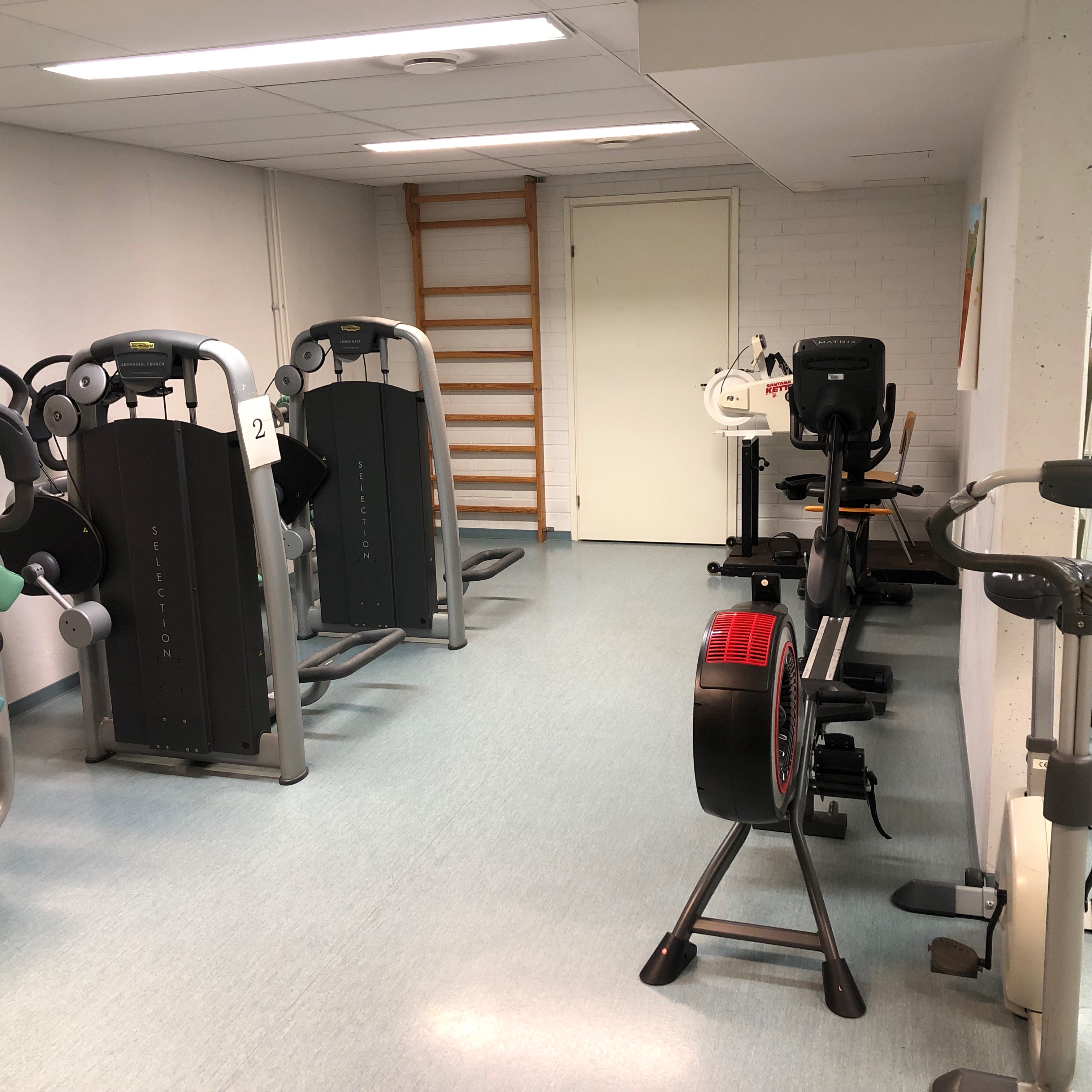 Kuntosali, jossa kaksi kuntosalilaitetta, soutulaite ja kuntopyörä valkoisella seinällä