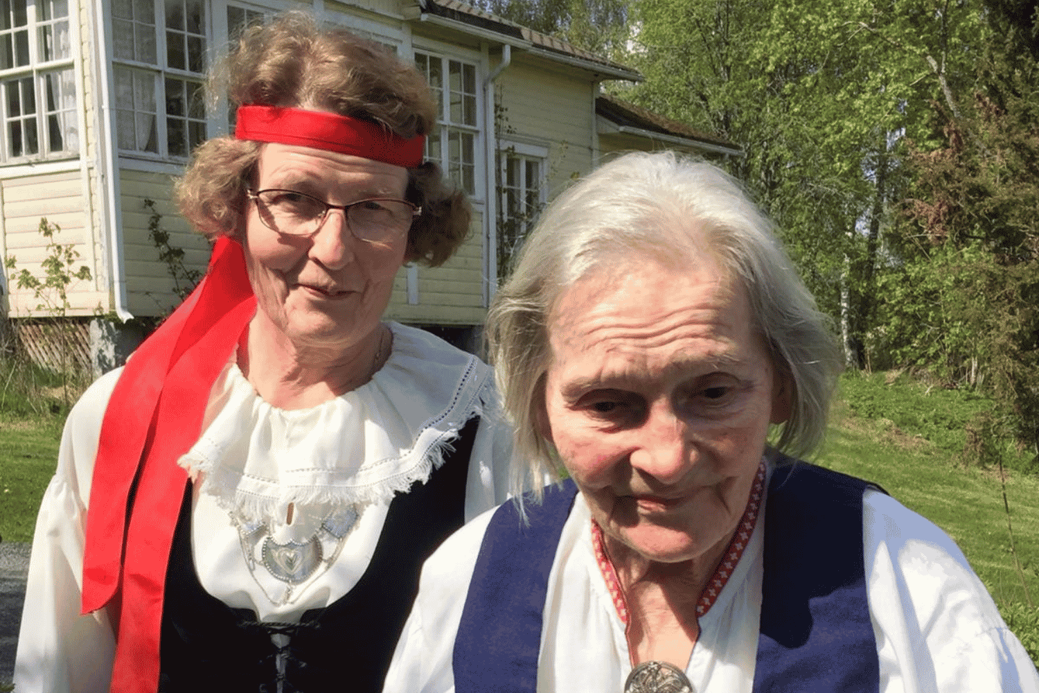 L&auml;heinen ja asukas seisovat ulkona keltaisen puutalon edess&auml;. Molemmilla on kansallispuvut. L&auml;heisell&auml; on punainen nauha hiuksissa.