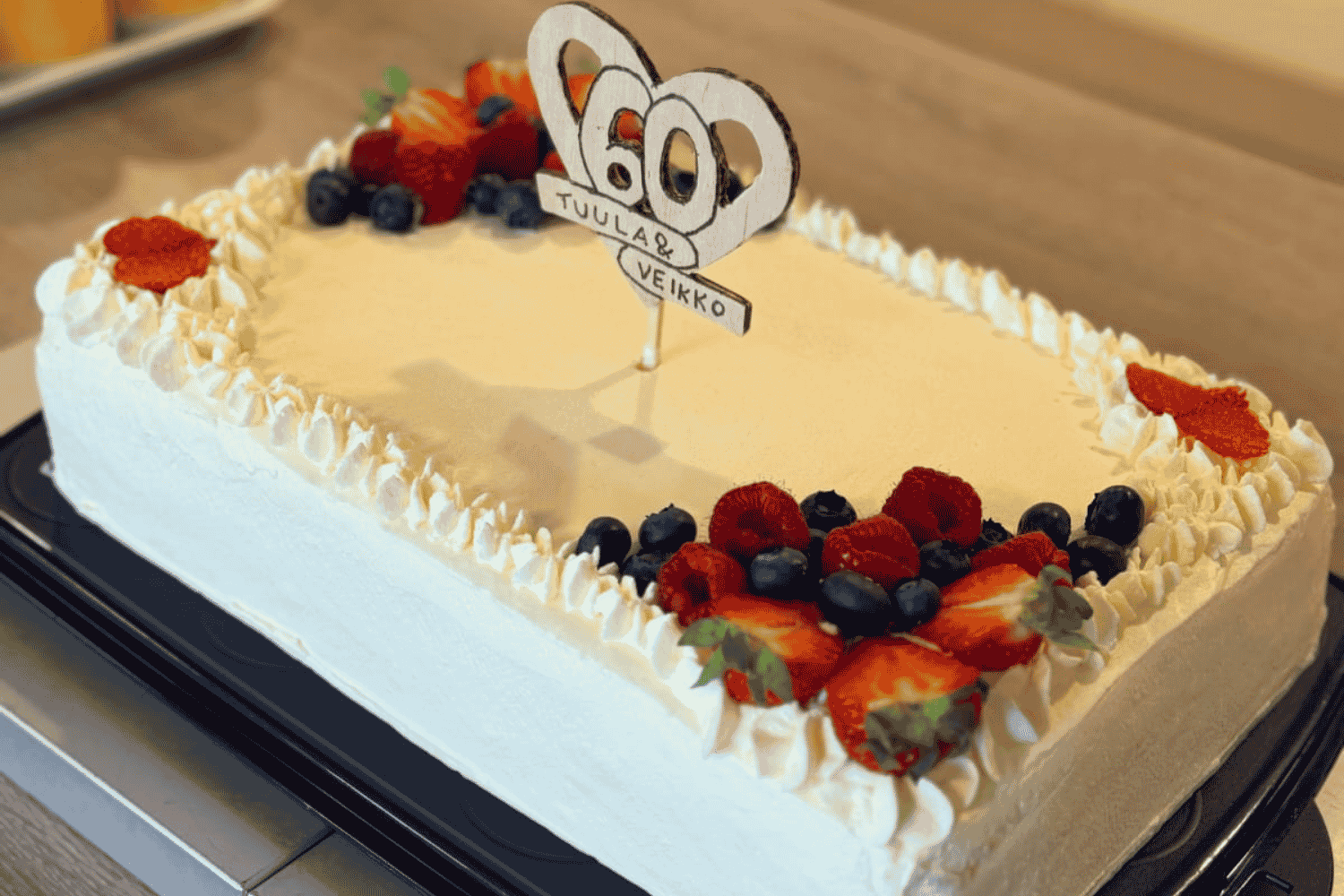 Juhlakakku, jonka p&auml;&auml;ll&auml; lukee "60, Tuula & Veikko". Kakun reunoilla on marjoja ja kermavaahtoa.