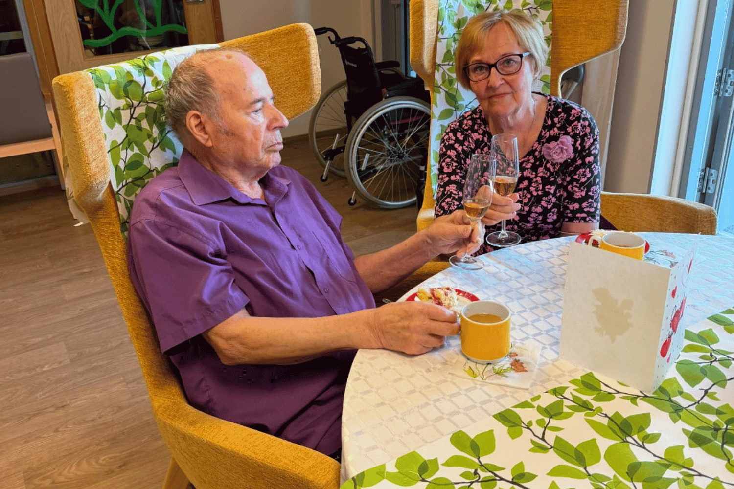 Veikko ja Tuula istuvat p&ouml;yd&auml;ss&auml; ja kohottavat maljan. P&ouml;yd&auml;ll&auml; on kahvia ja kakkua.