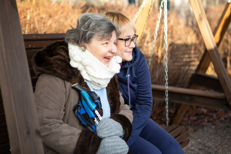 Hoivakodissa asuva nainen ja hoitaja istuvat hoivakodin pihalla pihakeinussa aurinkoisena loppusyksyn päivänä. Molemmat nauravat