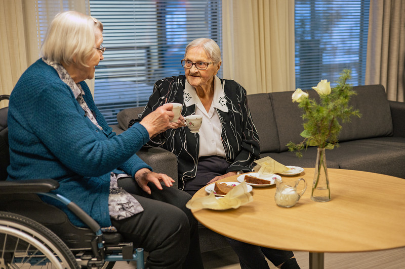 Kaksi iäkästä naista nauttimassa kahvia olohuoneessa pöydän ääressä, jossa on tarjoiltuna leivonnaisia ja kukkia maljakossa