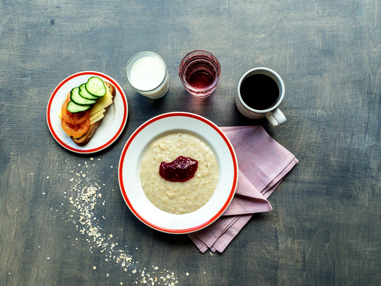 Aamupala katettuna harmaalle pöydälle, jossa on puuroa hillosilmällä, kahvia, mehua sekä leipää, jonka päällä on juustoa, kurkkua ja tomaattia